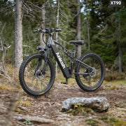 A black and dark gray Anish electric mountain bike with knobby tires is parked on a dirt trail in a forest setting. The brand name 'Anish' is clearly visible on the frame, and a model label 'XT90' is shown in the top right corner.