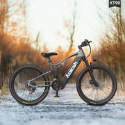 A dark gray Anlesh electric fat tire bike is parked on a snowy path outdoors with a blurred forest background. The bike features a matte dark gray frame with the 'Anlesh' brand name in white, black components, and knobby tires.