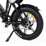 Close-up rear view of a black electric bicycle with a thick knobby tire, black fender, disc brake, and kickstand on a white studio background.