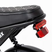 Close-up of the rear section of a black electric bicycle, showing the textured saddle, red rear light assembly, and part of the frame against a white background.