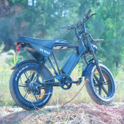 A matte black electric bicycle with fat tires, standing on a dirt path outdoors with a blurred green forest background. The bike features a step-through frame, a large battery mounted on the frame, and the text 'V6 Max' on the side panel.