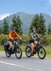 Two people riding electric bicycles on a paved road with a mountain and trees in the background. The person on the left wears an orange long-sleeve top and khaki shorts, while the person on the right wears an olive green long-sleeve top and black shorts.