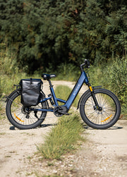 A blue step-through electric bicycle with a black rear pannier bag, parked on a dirt path outdoors with green trees and grass in the background.