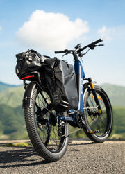 A blue electric bicycle with black pannier bags and a rear rack, parked on an asphalt road with a scenic mountain background under a blue sky.