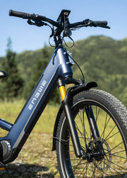 Close-up of a blue EnGUE electric bicycle with a front suspension fork, fender, and knobby tire, set against a blurred outdoor background of green hills and blue sky.