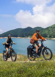 Two people riding electric bicycles on a grassy path next to a scenic lake with green hills in the background. The rider on the right wears an orange jacket and black helmet, while the rider on the left wears a green jacket and black helmet.