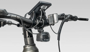 A close-up of a bicycle handlebar setup with a mounted black device, a small front light, a coiled cable, and a camera attached to the handlebar stem against a light gray background.