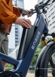 Close-up of a navy blue Engue electric bicycle with a person in an orange jacket touching the battery pack, set against a blurred city background.