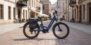 A blue GRIZZ electric cargo bike with a black rear pannier bag is parked on a cobblestone street in a European town.