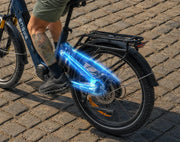 A close-up outdoor shot of a blue Enigma electric bicycle on a cobblestone surface, showing the rear wheel, rack, and a glowing blue graphic overlay highlighting the drivetrain area. A person's leg and foot are visible on the bike.
