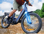 A close-up outdoor shot of a person riding an ENGLUE electric bicycle on a rocky dirt path under a cloudy sky. The bike frame is dark blue, the front fork is bright blue, and the rider is wearing khaki shorts and black shoes.