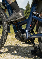 Close-up outdoor shot of a person riding an electric bicycle, showing the foot on the pedal, blue frame, motor, chainring, tire, and gravel path with trees in the background.