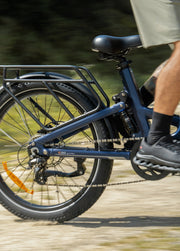Close-up side view of a blue electric cargo bicycle in motion, showing the rear wheel, rack, chainring, and part of a rider's leg and shoe on a blurred outdoor path.