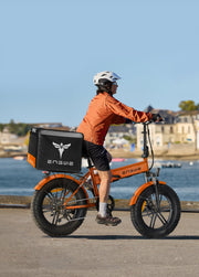 Person riding an orange electric bike with a black cargo box along a waterfront.