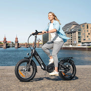 Woman riding an electric bike by a waterfront with buildings in the background