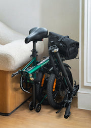 Foldable bicycle with a black bag on a wooden floor against a white wall.
