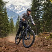 A man riding a black mountain bike on a dirt trail in a forest, with snow-capped mountains in the background. He is wearing a gray long-sleeve top, black pants, and a black helmet.